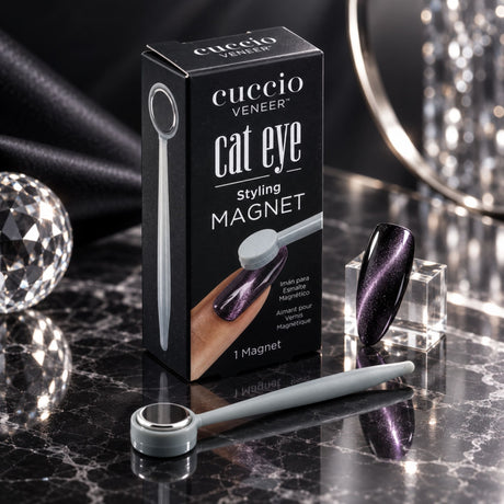 Cuccio Veneer Cat Eye Styling Magnet set on a polished black marble surface with reflective crystal props. The black retail box is centered, with a small light gray magnet and applicator tool placed in front, and a dark iridescent nail sample showing the cat-eye effect to the right.